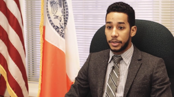 NYC Council Member Antonio Reynoso sitting at his desk, with the American and NYC City flags to his right.